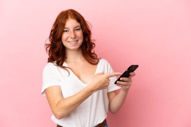 Genç kızılımsı kadın pembe arka planda izole edilmiş ve cep telefonu kullanıyor.