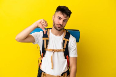 Sarı arka planda izole edilmiş büyük sırt çantalı genç bir beyaz adam başparmağını olumsuz ifade ile aşağı gösteriyor.