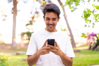Parktaki genç beyaz adam cep telefonuyla mesaj ya da e-posta yolluyor.