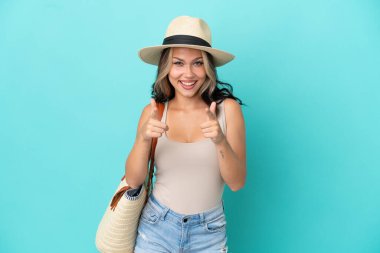 Pamel ve plaj çantalı genç Rus kız mavi arka planda izole edilmiş, ön tarafı işaret ediyor ve gülümsüyor.