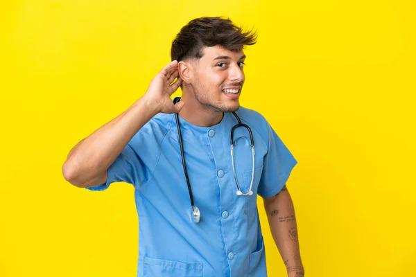 若い外科医の医師男は耳に手を置くことによって何かを聞いて黄色の背景に隔離 — ストック写真
