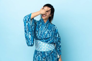 İzole edilmiş mavi arka planda kimono giyen genç bir kadın gözleri kaplıyor ve gülümsüyor.