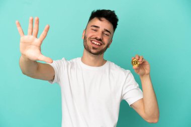 Elinde mavi arka planda izole edilmiş bir Bitcoin tutan genç adam parmaklarıyla beşi sayıyor