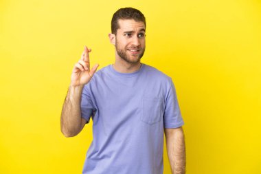 Sarışın yakışıklı adam izole edilmiş sarı arka planda parmaklarını çapraz yapıp en iyisini diliyor.