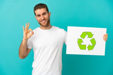 Yakışıklı, mavi arka planda, elinde geri dönüşüm simgesi olan bir pankart tutan ve zaferi kutlayan sarışın bir adam.