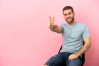 İzole edilmiş pembe arka planda oturan genç adam gülümsüyor ve zafer işareti gösteriyor.
