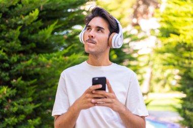 Parktaki genç beyaz adam cep telefonuyla müzik dinliyor ve düşünüyor.