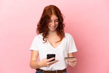 Pembe arka planda izole edilmiş genç bir kadın cep telefonuyla kredi kartıyla alışveriş yapıyor.