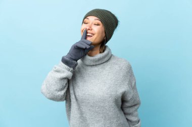 Mavi arka planda izole edilmiş kış şapkalı genç Uruguaylı kız sessiz jest yapıyor.