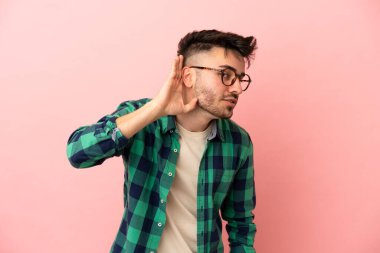 Pembe arka planda izole edilmiş genç beyaz adam elini kulağına koyarak bir şeyler dinliyor.