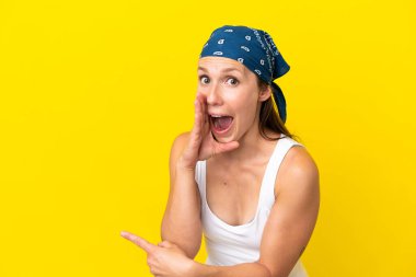 Sarı arka planda izole edilmiş genç bir İngiliz kadın bir ürün sunmak ve bir şeyler fısıldamak için kenarı işaret ediyor.