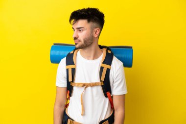 Sarı arka planda izole edilmiş büyük bir sırt çantası olan genç bir beyaz adam yan tarafa bakıyor.