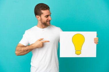 Sarışın, soyutlanmış, mavi arka planda, elinde ampul simgesi olan bir levha tutan yakışıklı bir adam.