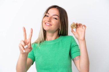 Beyaz arka planda izole edilmiş Bitcoin tutan genç Romen kadın gülümsüyor ve zafer işareti gösteriyor
