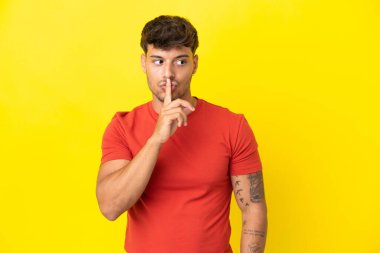 Sarı arka planda izole edilmiş beyaz, yakışıklı genç bir adam sessiz bir jest yapıyor. Parmağını ağzına sokuyor.
