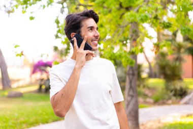 Parkta genç beyaz bir adam cep telefonuyla biriyle sohbet ediyor.