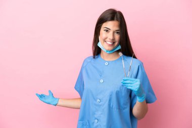 Pembe arka planda izole edilmiş aletleri tutan dişçi kadın davet edilmek için ellerini yana uzatıyor.