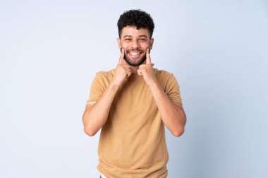 Faslı genç bir adam mavi arka planda izole edilmiş mutlu ve hoş bir ifadeyle gülümsüyor.