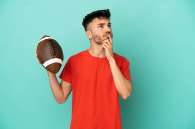 Rugby oynayan Kafkasyalı genç adam mavi arka planda izole edilmiş, yukarı bakarken şüpheleri var.