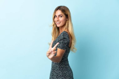 Genç sarışın Uruguaylı mavi arka planda izole edilmiş gülümsüyor ve zafer işareti gösteriyor.