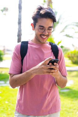 Parkta genç beyaz bir adam, mutlu bir ifadeyle cep telefonu kullanıyor.