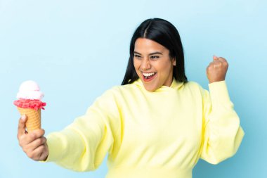 Kornet dondurmalı genç Kolombiyalı kadın mavi arka planda izole bir şekilde zaferi kutluyor.