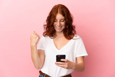 Pembe arka planda izole edilmiş genç bir kadın cep telefonu kullanıyor ve zafer jesti yapıyor.