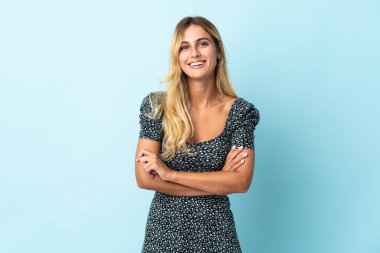 Genç sarışın Uruguaylı, mavi arka planda izole edilmiş. Kollarını ön cephede çapraz tutuyor.