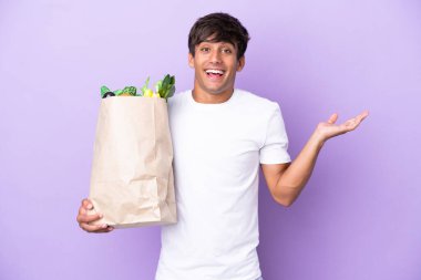 Elinde bir alışveriş çantası tutan genç adam mor arka planda izole edilmiş ve şok olmuş bir yüz ifadesi var.