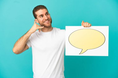 Yakışıklı sarışın, izole edilmiş mavi arkaplan üzerinde konuşma baloncuğu ikonu olan bir levha tutuyor ve telefon jesti yapıyor.