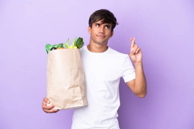 Elinde bir alışveriş çantası tutan genç adam mor arka planda izole edilmiş ve şans diliyor.