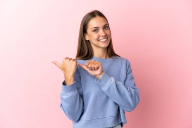 Soyutlanmış pembe arka planı olan genç bir kadın bir ürünü sunmak için kenarı işaret ediyor.