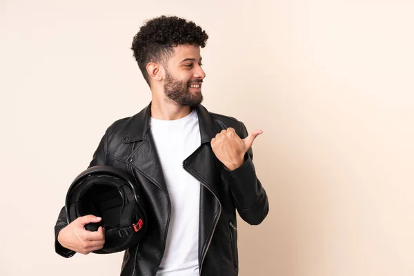 Young Moroccan man with a motorcycle helmet isolated on beige background pointing to the side to present a product