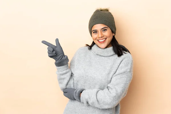 年轻的哥伦比亚女孩 戴着冬帽 被米色背景隔离 手指指向侧面 — 图库照片