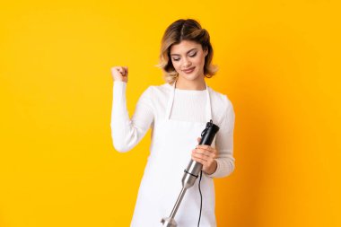 Sarı arka planda izole edilmiş el blenderi kullanan kız zaferi kutluyor.