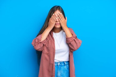 Mavi arka planda izole edilmiş genç beyaz kadın gözlerini elleriyle kapatıyor.