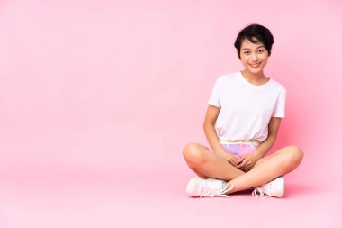 Kısa saçlı genç Vietnamlı kadın pembe arka planda izole bir şekilde oturmuş gülüyor.