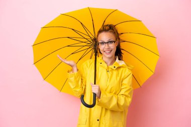 Yağmura dayanıklı paltosu ve şemsiyesi olan genç Rus kız pembe arka planda, şok olmuş yüz ifadesi ile izole edilmiş.