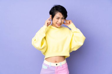 Mor arka planı yüzünden kısa saçlı genç Vietnamlı kadın hayal kırıklığına uğramış ve kulaklarını kapatmış.