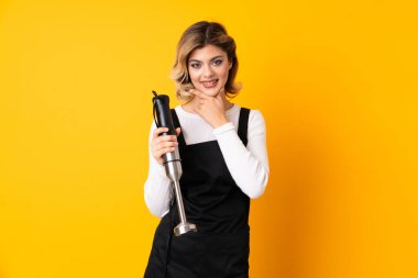 Sarı arka planda izole edilmiş el blenderi kullanan kız yana bakıyor ve gülümsüyor.