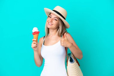 Elinde dondurma ve plaj çantası olan genç Romen kadın mavi arka planda izole edilmiş, başparmakları havada, çünkü iyi bir şey olmuş.