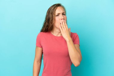 Genç beyaz kadın mavi arka planda izole edilmiş esniyor ve geniş bir ağzı eliyle kapatıyor.