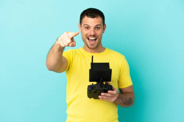 İzole edilmiş mavi arka plan üzerinde uzaktan kumanda tutan Brezilyalı adam şaşırmış ve ön tarafı işaret ediyor.