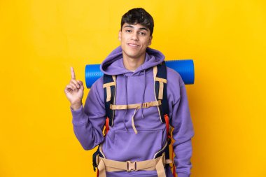 Sarı arka plan üzerinde büyük bir sırt çantası olan genç dağcı en iyinin işareti olarak parmağını kaldırıyor.