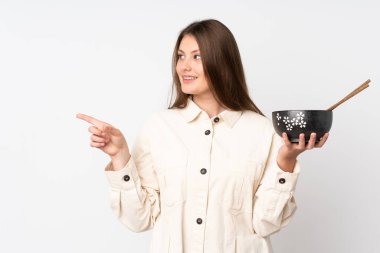 Beyaz arka planda izole edilmiş genç beyaz bir kız elinde çubuklarla elinde bir kase erişte tutarken bir ürünü sunmak için kenarı işaret ediyor.