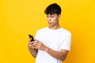 Sarı arka plan izole edilmiş genç adam kameraya bakarken şaşırmış bir ifadeyle cep telefonunu kullanıyor.