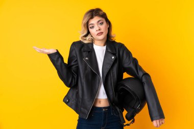 Sarı arka planda izole edilmiş motosiklet kaskı tutan genç bir kadın. Kafası karışmış bir yüz ifadesi var.