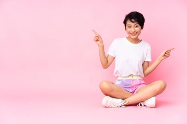 Kısa saçlı genç Vietnamlı kadın, pembe arka planda izole edilmiş bir şekilde yerde oturuyor. Yan tarafları işaret ediyor ve mutlu.
