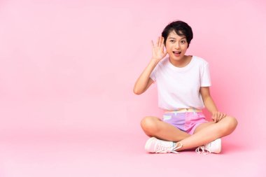 Kısa saçlı genç Vietnamlı kadın, pembe arka planda izole edilmiş bir şekilde yerde oturuyor ve elini kulağına koyarak bir şeyler dinliyor.