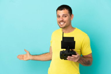 İzole edilmiş mavi arka plan üzerinde uzaktan kumandayı elinde tutan Brezilyalı bir adam davet için elini yana uzatıyor.